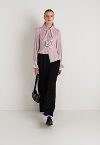 Blusa rosa chiaro con volant davanti e colletto, maniche lunghe con polsini, abbinata a pantaloni neri e borsa nera. Tessuto morbido, vestibilità su misura.