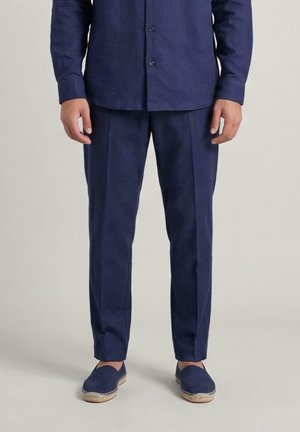 Uomo che indossa una camicia blu navy abbottonata, pantaloni su misura abbinati e espadrillas blu navy, in piedi su uno sfondo chiaro.