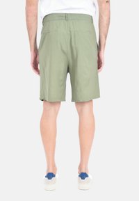Pantaloni shorts verde oliva con vita elasticizzata e due tasche posteriori. Tessuto morbido, lunghezza sopra il ginocchio, abbinati a sneakers bianche e blu.