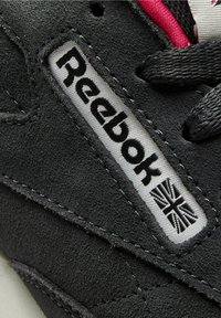 Détail d'une basket Reebok en daim noir avec une étiquette blanche indiquant « Reebok » et un drapeau noir de l’Union Jack près des lacets noirs avec une bordure rouge.