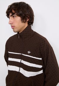 Καφέ τζάκετ με φερμουάρ που διαθέτει υφή και τρεις λευκές οριζόντιες ρίγες; λογότυπο Adidas στο αριστερό στήθος. Ψηλός γιακάς και άνετη εφαρμογή.