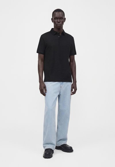 Homme debout portant un polo noir à manches courtes, un jean bleu clair ample et des chaussures noires sur un fond blanc uni.