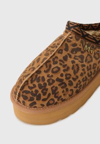 Bruine pantoffels met een luipaardprint, gemaakt van zacht suede materiaal, met een gewatteerde binnenkant, rubberen zool en een decoratieve kraag met zwart patroon.