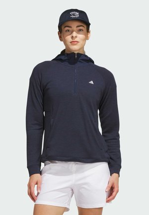 Kvinde iført en marineblå halvlynlåshættetrøje, hvide shorts og en marineblå Adidas kasket, stående mod en ensfarvet lys baggrund.