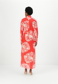 Rochie roșie cu mâneci lungi, cu mari modele de scoici albe, cu nasturi la spate și o silueta fluidă. Țesătură netedă cu o textură ușoară.