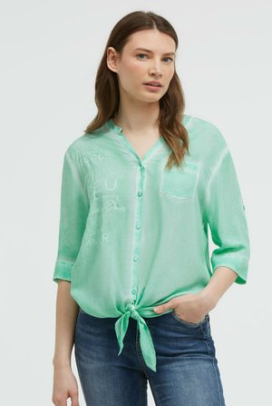 Junge Frau mit braunen Haaren, die eine hellgrüne, geknöpfte Bluse trägt, die in der Taille gebunden ist, und blaue Jeans, steht mit einer Hand in der Tasche.