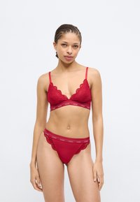 Rotes Spitzen-Bralette mit Dreiecksschalen und verstellbaren Trägern; passender roter Tanga mit Spitzenrändern und Logoband; glatte Stoffstruktur.