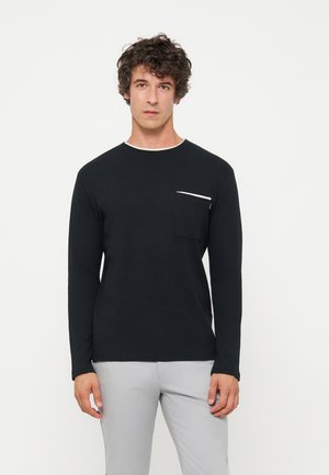 Homme aux cheveux bouclés portant une chemise noire à manches longues avec une poche poitrine et un pantalon gris clair, debout devant un fond uni.