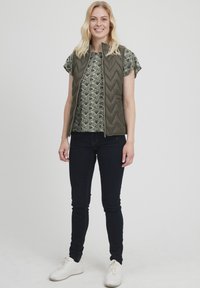 Zöld steppelt mellény magas gallérral, mintás felsőre párosítva, sötét, slim fit farmernadrággal és fehér sportcipővel egy laza megjelenésért.