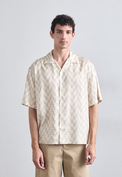Chemise à manches courtes, beige, avec un motif géométrique texturé. Dotée d'un col évasé et de boutons blancs, associée à un short beige clair.