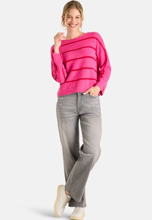 Woman smiling wearing pink striped sweater, gray wide-leg jeans with "READY SET GLOW" text, and white sneakers, standing with one hand in pocket.