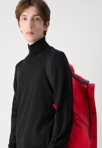 Ung person med kort brunt hår iført en sort turtleneck og bærende på en rød dunjakke over den ene skulder mod en ensfarvet baggrund.
