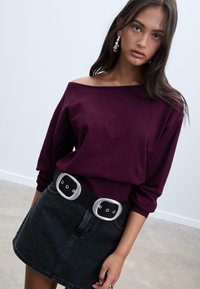 Un pull maroon à épaules dénudées associé à une jupe en denim noire. La jupe est ornée d'une large ceinture argentée avec un design à double boucle.