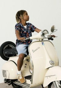 Vespa scooter color crema con accenti in cromo, dotata di un sedile nero. Bambino in camicia floreale e pantaloni corti di denim seduto sulla scooter.