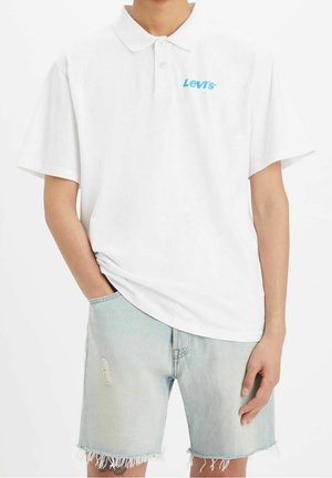 Personne portant un polo blanc à manches courtes Levi's avec un short en denim déchiré bleu clair, debout avec une main dans la poche.