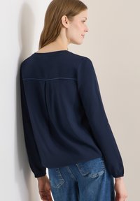 Femme vêtue d'un chemisier bleu marine à manches longues avec un détail plissé, debout de côté contre un mur clair, portant un jean bleu.