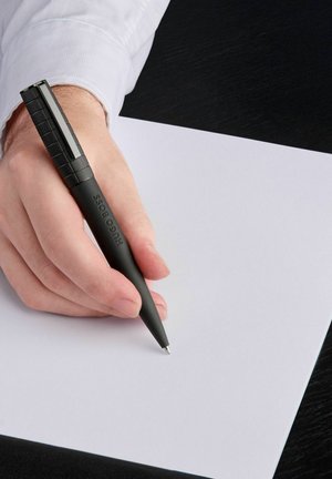 Zwarte balpen vastgehouden in een hand, met een ribbelige grip, zilveren clip en "Hugo Boss" gegraveerd. Schrijvend op een wit vel papier.