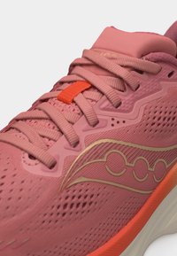 Ροζ αθλητικό παπούτσι με υφή υφάσματος, ροζ κορδόνια, πορτοκαλί ταμπελάκι κορδονιών και χρυσό λογότυπο στο πλάι, με λευκή και πορτοκαλί σόλα.