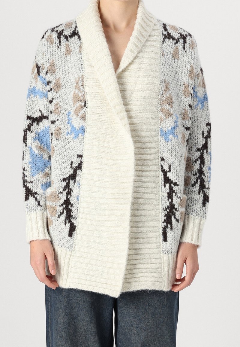 Personne portant un cardigan épais à ouverture avant de couleur crème avec un col côtelé et des motifs floraux bleus, beige et noirs sur les manches et les côtés.