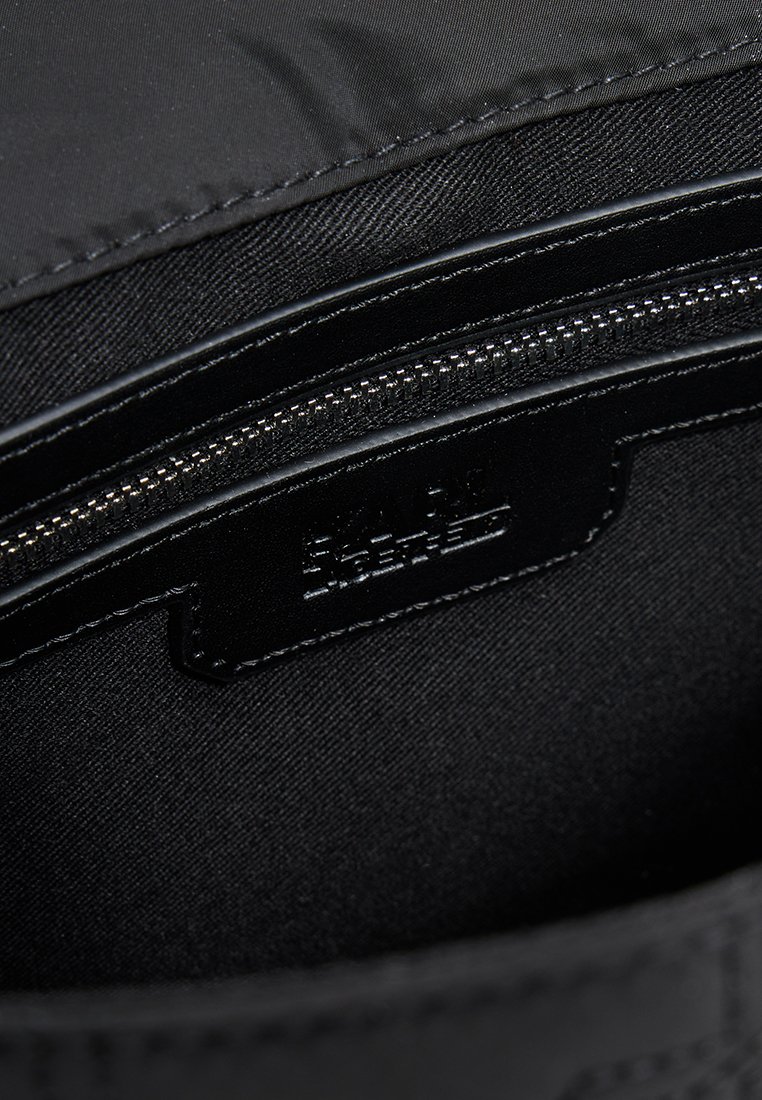 Intérieur d'un sac en tissu noir présentant une poche zippée, une doublure synthétique lisse et une étiquette de marque embossée avec de subtils détails de couture.