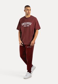 Bordeauxkleurige set bestaande uit een losse t-shirt met witte tekst "SMILODOX ATHL. WEAR" en joggingbroek met een logo op de dij. Witte sneakers.