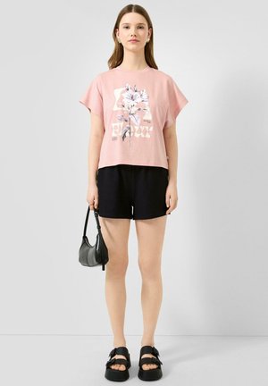 Femme portant un T-shirt rose avec un imprimé floral, un short noir, des sandales noires et tenant un petit sac à main noir, debout devant un fond uni.