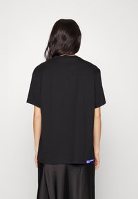 Camiseta de algodón negra con un corte relajado, mangas cortas y cuello redondo. Presenta una pequeña etiqueta de logo azul en el dobladillo del lado izquierdo.