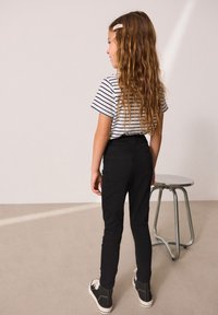Pantalones negros ajustados con una cinturilla elástica, combinados con una camisa de rayas de manga corta. Pelo largo y ondulado y zapatillas negras visibles.