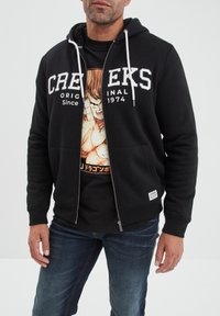 Sudadera negra con cremallera y texto blanco "CREWERS" junto con el logo; forro interior suave, capucha ajustable con cordón, bolsillos frontales, superpuesta sobre una camiseta gráfica.