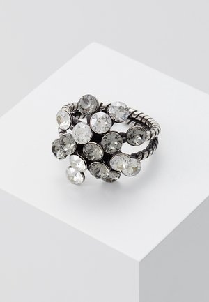 Bague en argent avec bande enroulée ornée de pierres précieuses rondes claires regroupées, disposées en design floral, posée sur un piédestal blanc.