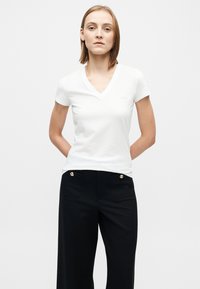 Λευκό t-shirt με V λαιμόκοψη από απαλό ύφασμα, συνδυασμένο με μαύρο παντελόνι που έχει χρυσά κουμπιά και μπροστινές τσέπες· απλός, μοντέρνος σχεδιασμός.