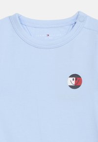 Φωτεινό γαλάζιο βρεφικό κορμάκι με στρογγυλή λαιμόκοψη, κουμπί πίσω στον ώμο και μικρό στρογγυλό λογότυπο με χαμογελαστό πρόσωπο σε μαύρο, άσπρο και κόκκινο.