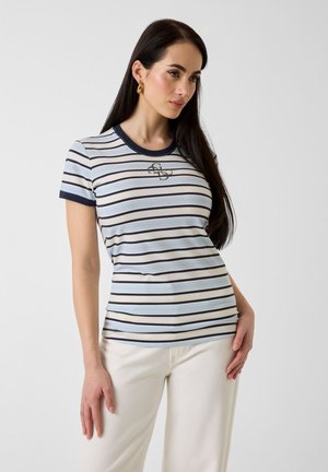 Femme aux longs cheveux foncés portant un t-shirt ajusté à manches courtes rayé avec des accents bleu marine et un pantalon écru, posant devant un fond uni.