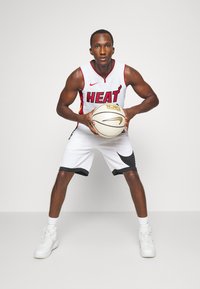 Basketbola spēlētājs baltā krekliņā ar uzrakstu "HEAT" sarkanā krāsā, pelēkās šortos, turot baltu basketbola bumbu ar zelta akcentiem, apģērbts baltās sporta apavās.