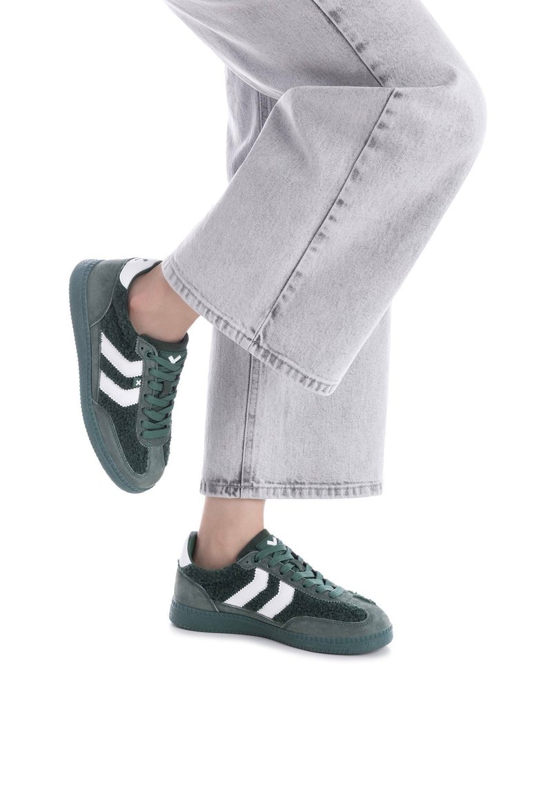 Zapatillas verdes con rayas blancas, materiales de textil y ante, acentos texturizados, diseño con cordones, combinadas con jeans de pierna ancha gris claro.