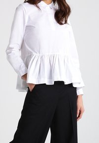 Blusa bianca a maniche lunghe con un corpino aderente e un orlo peplum svasato. Presenta un colletto e una discreta fila di bottoni frontali.