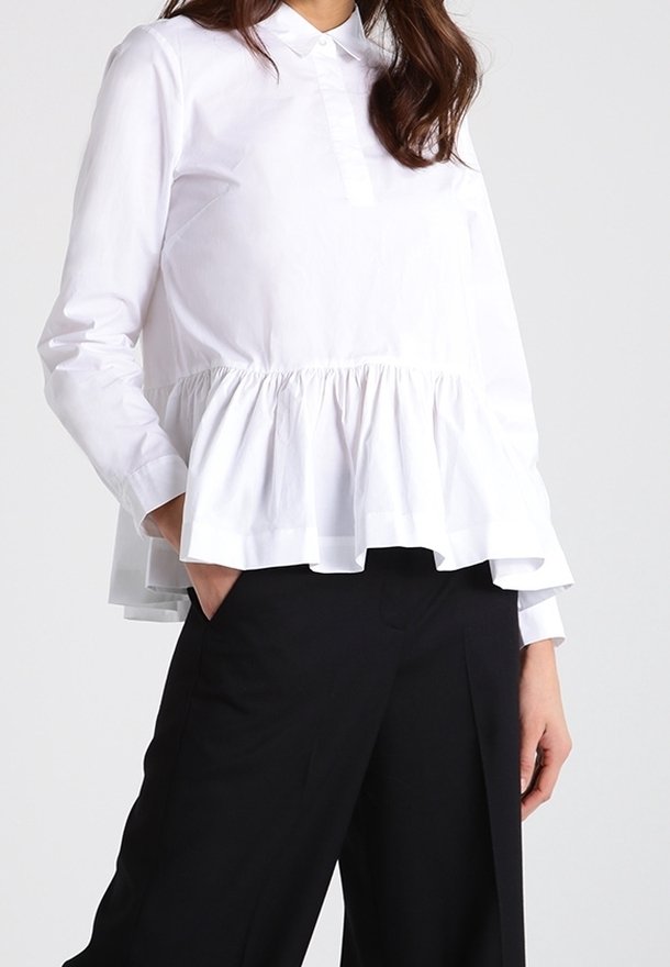 Blusa bianca a maniche lunghe con un corpino aderente e un orlo peplum svasato. Presenta un colletto e una discreta fila di bottoni frontali.