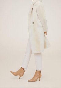 Witte nepleren bontjas met ronde kraag en knoopdetails, gecombineerd met crèmekleurige skinny jeans en beige enkellaarsjes met spitse neuzen en uitlopende hielen.