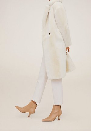 Witte nepleren bontjas met ronde kraag en knoopdetails, gecombineerd met crèmekleurige skinny jeans en beige enkellaarsjes met spitse neuzen en uitlopende hielen.