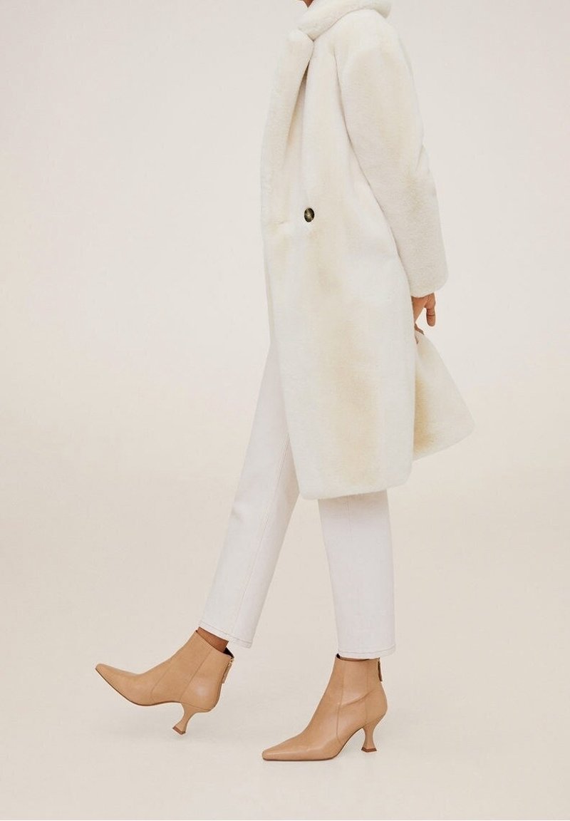 Witte nepleren bontjas met ronde kraag en knoopdetails, gecombineerd met crèmekleurige skinny jeans en beige enkellaarsjes met spitse neuzen en uitlopende hielen.