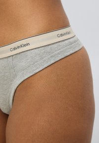 Γκρι βαμβακερό στρινγκ με μπεζ ελαστική μέση που φέρει μαύρο κείμενο "Calvin Klein", αναδεικνύοντας έναν ομαλό, εφαρμοστό σχεδιασμό.