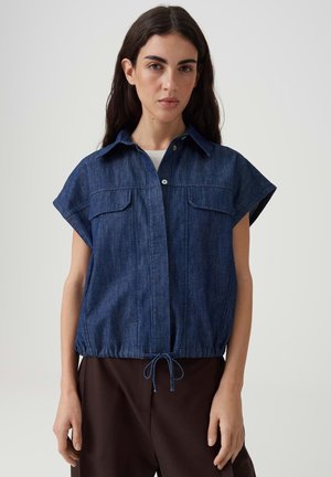 Femme aux longs cheveux foncés portant une veste en denim foncé à manches courtes avec poches avant et cordon de serrage à l'ourlet, debout devant un fond uni.