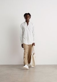 Hvid skjorte med knapper, beige bukser og hvide sneakers. Modellen holder en beige tote taske. Simpelt design, glat tekstur, minimale accenter.
