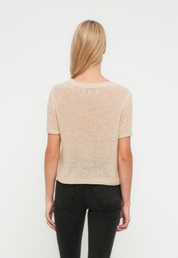 Bež pletena majica kratkih rukava s opuštenim krojem, okruglim izrezom i teksturiranim završetkom. Pogled s leđa pokazuje ravni rub.