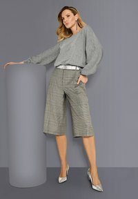 Frau in einem grauen Pullover mit Ballonärmeln, karierterweit geschnittener, halbhoher Hose und silbernen High Heels, die sich an einem grauen zylindrischen Sockel lehnt.