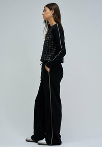 Femme portant un haut à manches longues à carreaux noirs avec des passepoils blancs aux manches, associé à un pantalon large noir avec des poches latérales et des baskets blanches.