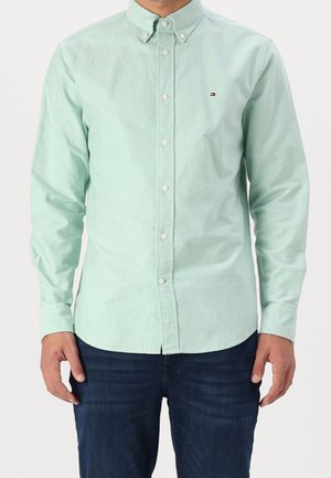 Homme portant une chemise vert clair à boutons avec manches longues et un jean bleu foncé, debout devant un fond uni.