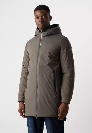 Hombre con chaqueta de invierno larga gris con capucha, cremallera frontal y bolsillos laterales con cremallera, de pie frente a fondo blanco.
