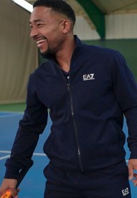 Giacca sportiva con zip di colore blu navy abbinata a pantaloni, caratterizzata da una texture liscia e logo EA7 sul petto e sulla gamba. Indossata in un contesto sportivo al chiuso.
