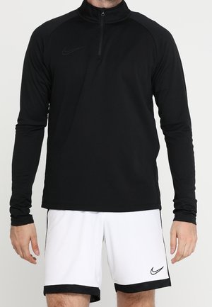 Mężczyzna noszący czarną bluzę z długim rękawem i półzamkiem oraz białe spodenki Nike z czarnym obszyciem i logo na lewej nodze.